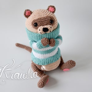 May include: A crocheted stuffed animal resembling a raccoon wearing a teal and white striped jumper. The animal has brown eyes, a brown nose, and pink ears.