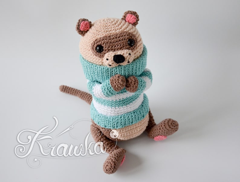May include: A crocheted stuffed animal resembling a raccoon wearing a teal and white striped jumper. The animal has brown eyes, a brown nose, and pink ears.