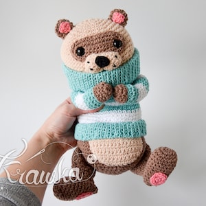 May include: A crocheted raccoon plush toy wearing a blue and white striped jumper. The toy has brown fur, black eyes, and pink ears and paws.