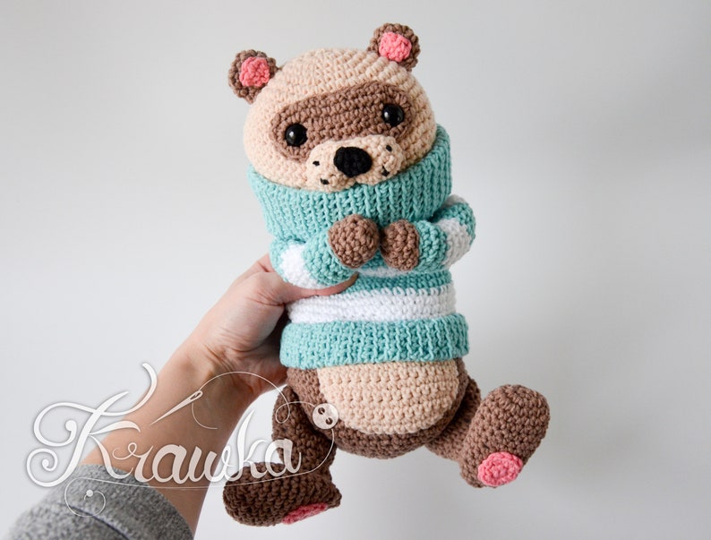May include: A crocheted raccoon plush toy wearing a blue and white striped jumper. The toy has brown fur, black eyes, and pink ears and paws.