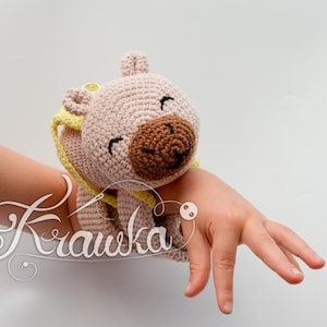 May include: A crocheted bear wrist rattle in light brown with a brown snout and black eyes. The rattle has yellow accents and is worn on a wrist. The word "Krawka" is written in cursive on the image.
