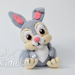 May include: A grey crocheted rabbit plush toy with pink ears, a white heart on its chest, and brown eyes. The toy is sitting on a white surface.