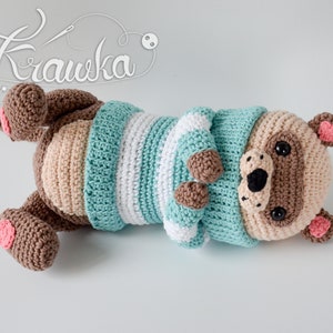 May include: A crocheted stuffed animal in the shape of a bear wearing a blue and white striped sweater. The bear has brown fur, a black nose, and pink ears.