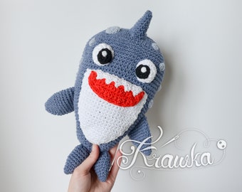 May include: A crocheted gray shark plush toy with white teeth and red mouth. The shark has black eyes and white spots.