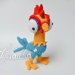 Puede incluir: Un juguete de gallo de crochet con una cresta roja, naranja y amarilla, ojos grandes blancos y un cuerpo azul. El gallo está de pie sobre una superficie blanca.