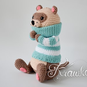 May include: A crocheted stuffed animal wearing a blue and white striped jumper and a brown scarf. The animal has brown fur, a pink nose, and pink ears.