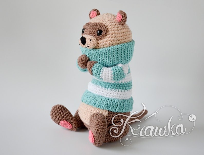 May include: A crocheted stuffed animal wearing a blue and white striped jumper and a brown scarf. The animal has brown fur, a pink nose, and pink ears.