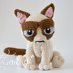 May include: A crocheted stuffed cat with brown and white fur, wearing brown sunglasses and a grumpy expression. The cat is sitting on a white surface.