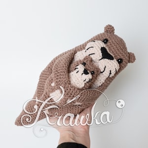 May include: A crocheted brown otter with a smaller otter nestled inside. The otters have black eyes and cream-colored faces and paws. The image includes the word "Krawka" in a cursive font.