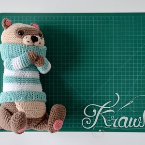 May include: A crocheted stuffed animal resembling a raccoon wearing a teal and white striped sweater. The animal is lying on a green cutting mat with the word "Krawka" written in white.
