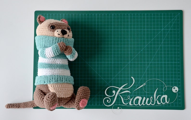 May include: A crocheted stuffed animal resembling a raccoon wearing a teal and white striped sweater. The animal is lying on a green cutting mat with the word "Krawka" written in white.