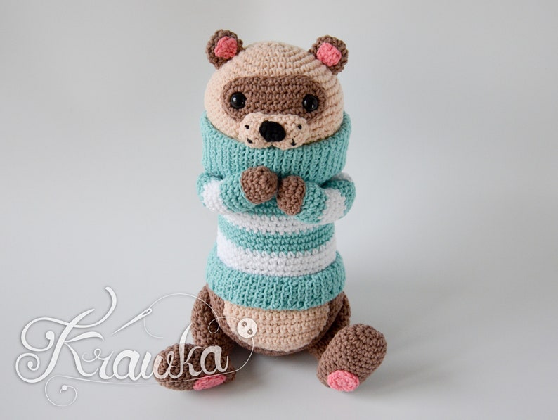 May include: A crocheted stuffed animal in the shape of a bear wearing a blue and white striped jumper. The bear has brown fur, a pink nose, and black eyes.