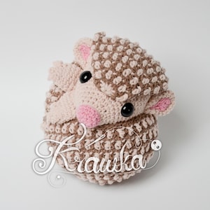May include: A crocheted hedgehog plush toy with brown and pink yarn. The hedgehog has black eyes and a pink nose.