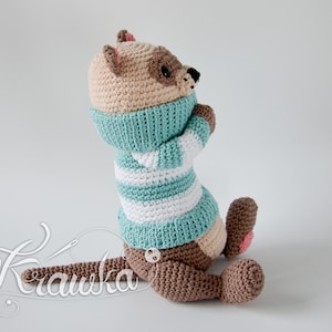 May include: A crocheted stuffed animal in the shape of an otter, made in brown and beige yarn, wearing a teal and white striped sweater. The otter is sitting on a white surface.