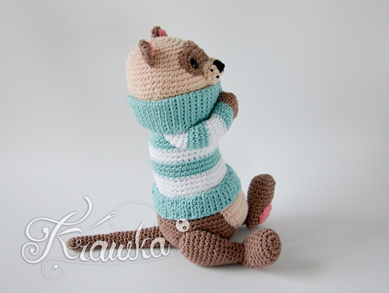 May include: A crocheted stuffed animal in the shape of an otter, made in brown and beige yarn, wearing a teal and white striped sweater. The otter is sitting on a white surface.