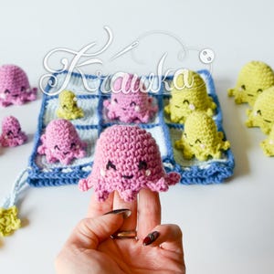 May include: Handmade crochet octopus toys in various sizes and colors. The octopuses are primarily pink and yellow, with smiling faces and small tentacles. Some are arranged on a blue and white crocheted square, while others are scattered around.