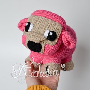 May include: Pink crocheted sheep plush toy with black eyes and a black nose. The sheep is holding a small white button in its mouth.