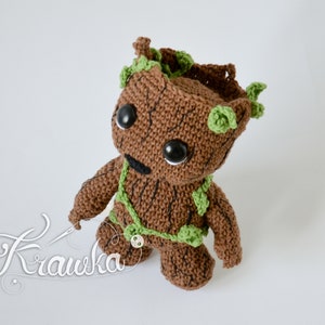Puede incluir: Un personaje de Groot de crochet marrón con hojas verdes y ojos negros. El personaje está de pie sobre un fondo blanco.