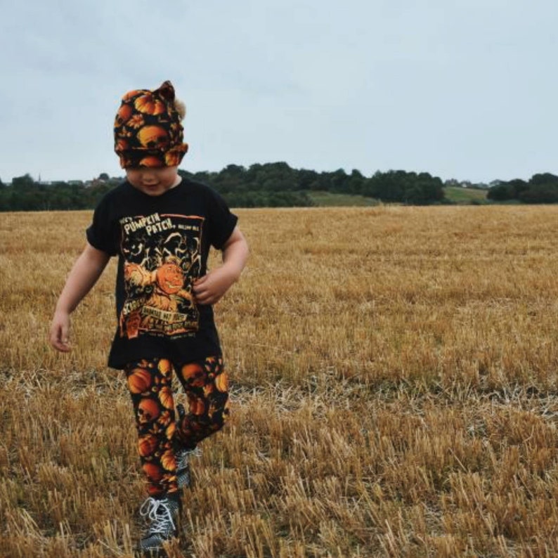 May include: A child wearing a black t-shirt with a pumpkin patch graphic and matching pumpkin print leggings. The child is wearing a pumpkin-themed beanie and is standing in a field of dry grass.