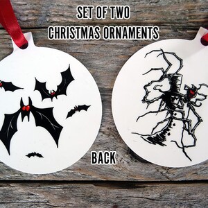 May include: Set of two white Christmas ornaments with red ribbon hangers. One ornament features black bats with red accents, the other a black and white illustration of a creature. Text on the ornaments reads "SET OF TWO CHRISTMAS ORNAMENTS" and "BACK".