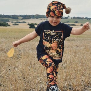 May include: A child wearing a black t-shirt with a pumpkin patch graphic and orange and black skull leggings. The child is wearing a black and orange skull beanie with a fuzzy pom pom. The child is holding a yellow leaf and running through a field of dry grass.