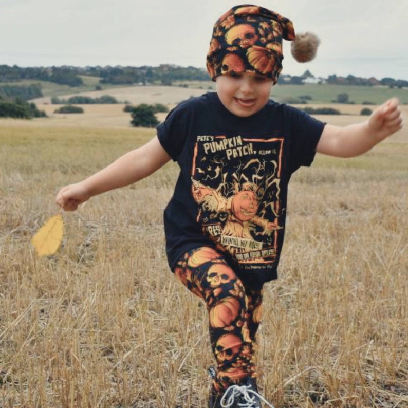May include: A child wearing a black t-shirt with a pumpkin patch graphic and orange and black skull leggings. The child is wearing a black and orange skull beanie with a fuzzy pom pom. The child is holding a yellow leaf and running through a field of dry grass.