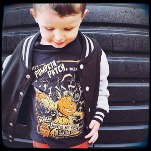 May include: A young boy wearing a black and white varsity jacket with a pumpkin patch themed t-shirt. The t-shirt features a cartoon pumpkin with the text "ETE'S PUMPKIN PATCH HOLLO SCARES! NOW WIN 400 HAUNTED HAY RIDES! DIP YOUR OWN POISON APPLE! WHAT ARE YOU SCARED OF?"