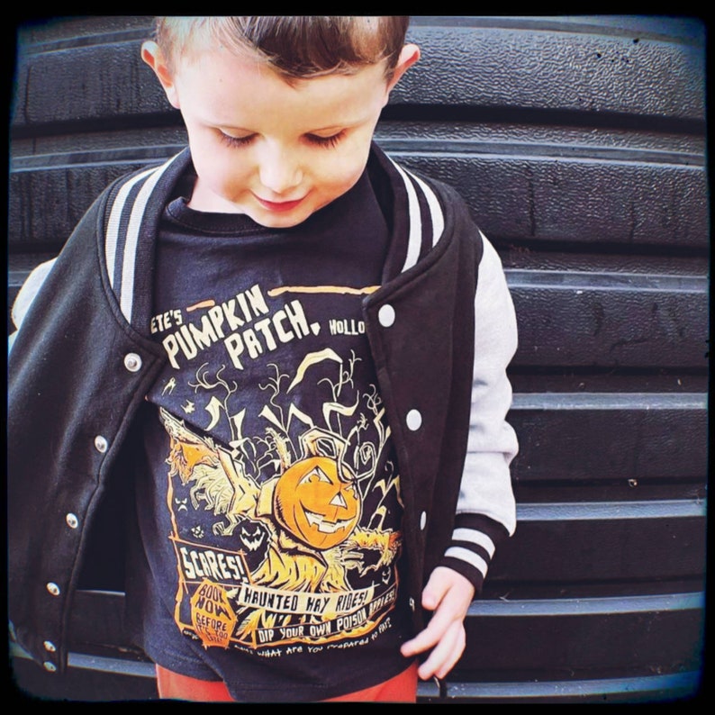 May include: A young boy wearing a black and white varsity jacket with a pumpkin patch themed t-shirt. The t-shirt features a cartoon pumpkin with the text "ETE'S PUMPKIN PATCH HOLLO SCARES! NOW WIN 400 HAUNTED HAY RIDES! DIP YOUR OWN POISON APPLE! WHAT ARE YOU SCARED OF?"