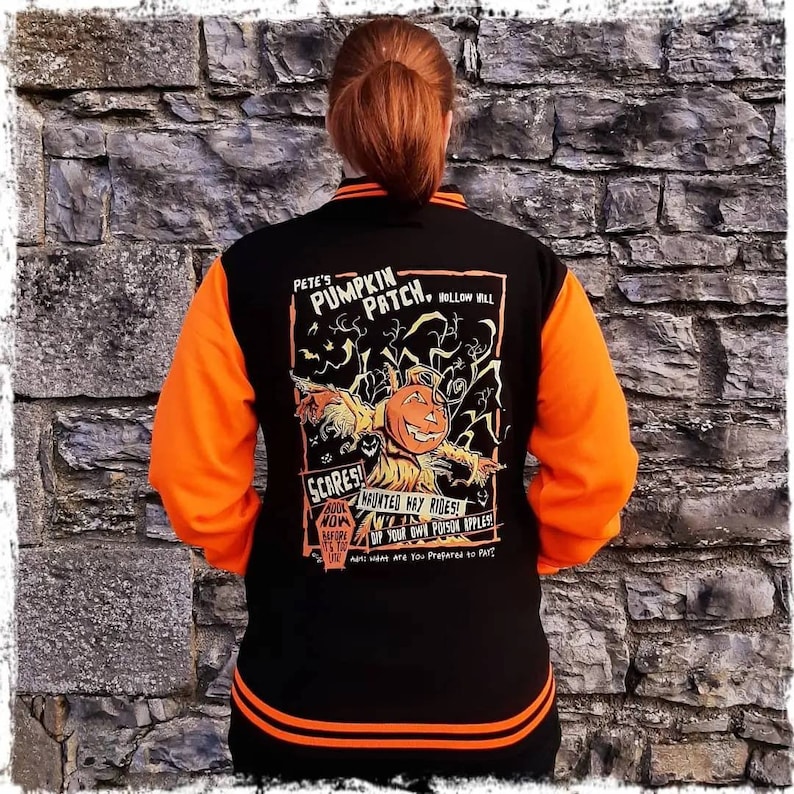 May include: Black and orange baseball jacket with a Halloween-themed design on the back. The design features a pumpkin patch with a scarecrow and the text "Pete's Pumpkin Patch, Hollow Hill".