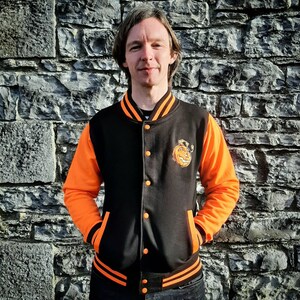 May include: A black and orange varsity jacket with a pumpkin face graphic on the left chest. The jacket has a zipper closure and ribbed cuffs and waistband.