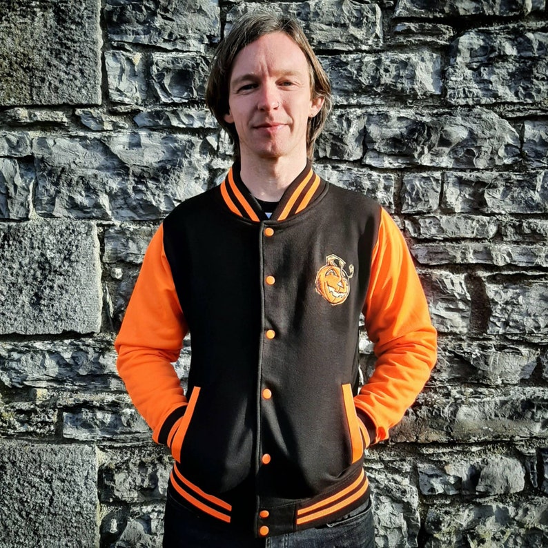 May include: A black and orange varsity jacket with a pumpkin face graphic on the left chest. The jacket has a zipper closure and ribbed cuffs and waistband.