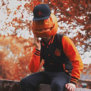 May include: A person wearing a black and orange varsity jacket with a pumpkin carved face as a hat. The person is sitting on a wall with a blurred background of fall foliage.