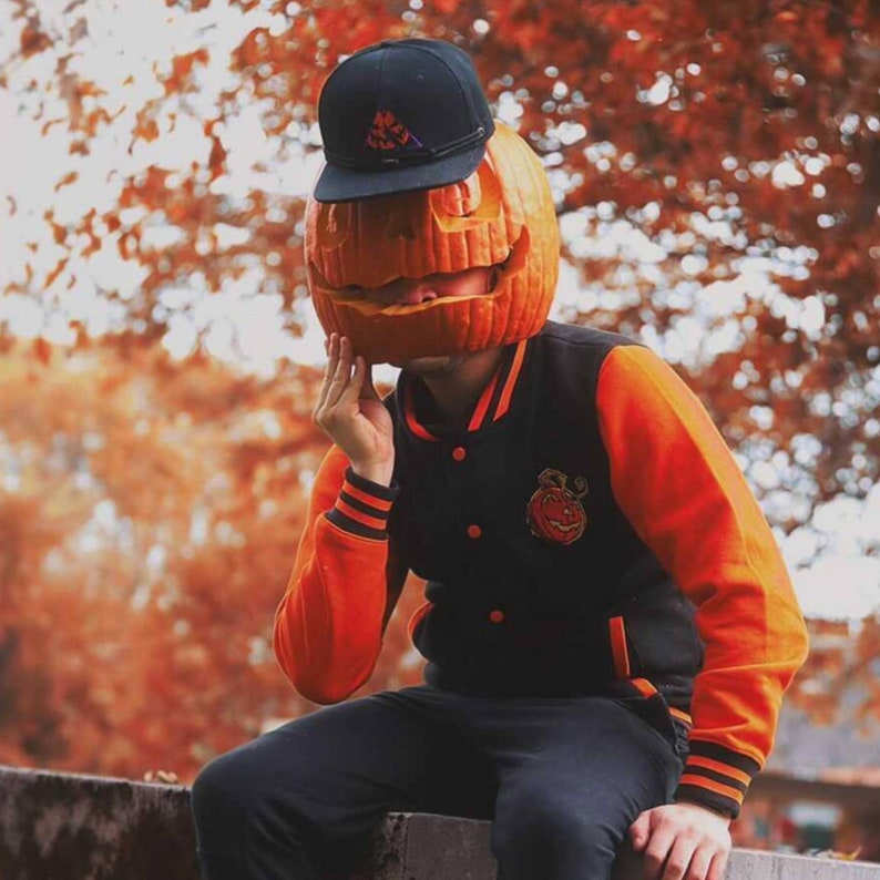May include: A person wearing a black and orange varsity jacket with a pumpkin carved face as a hat. The person is sitting on a wall with a blurred background of fall foliage.
