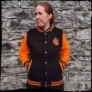 May include: A black and orange varsity jacket with a pumpkin face embroidered on the left chest. The jacket has orange sleeves and a black body with orange stripes on the cuffs and bottom.