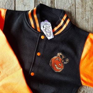 May include: Black and orange varsity jacket with an embroidered pumpkin on the front. The jacket has a black body with orange sleeves and a black collar with orange stripes. The tag on the jacket reads "Hollow Hill Apparel".