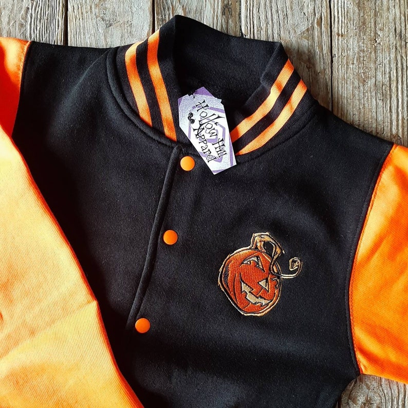 May include: Black and orange varsity jacket with an embroidered pumpkin on the front. The jacket has a black body with orange sleeves and a black collar with orange stripes. The tag on the jacket reads "Hollow Hill Apparel".
