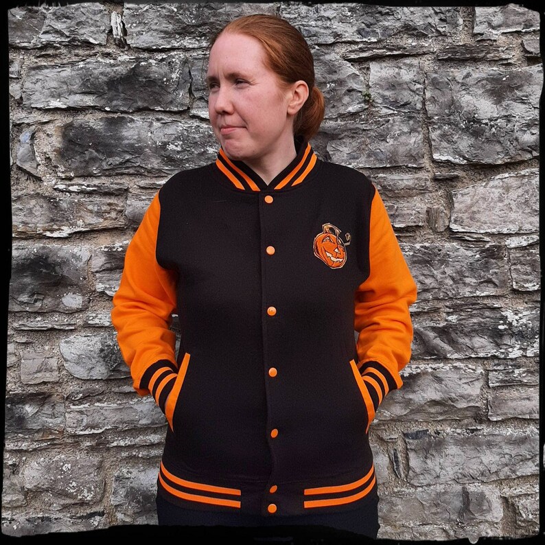 May include: A black and orange varsity jacket with a pumpkin face embroidered on the left chest. The jacket has orange sleeves and black body with orange stripes on the cuffs and bottom.