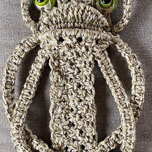 May include: A macrame wall hanging with a brown and white rope design. The hanging features two large green eyes and a bamboo hoop at the top.