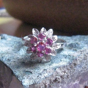 Op de afbeelding: Een zilveren ring met een cluster van roze edelstenen in een bloemvorm. De ring wordt getoond op een paarse amethistkristal.