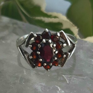 May include: A silver ring with a floral design of dark red gemstones. The central oval stone is surrounded by smaller, round stones. The ring's band has an openwork design on either side, adding to its decorative appeal.