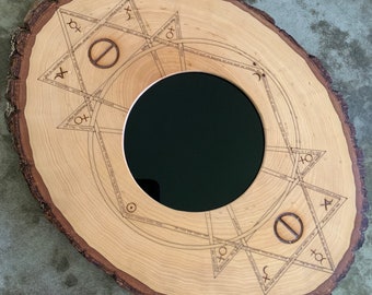 May include: A round wooden mirror with a natural edge. The light-colored wood is etched with geometric shapes and text around a central circular mirror. The mirror has a dark, reflective surface.