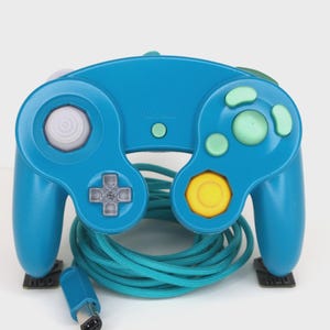 May include: A teal-colored video game controller with a variety of buttons in shades of gray, green, and yellow. The controller has a matching teal cord and connector. The controller is resting on a white surface.