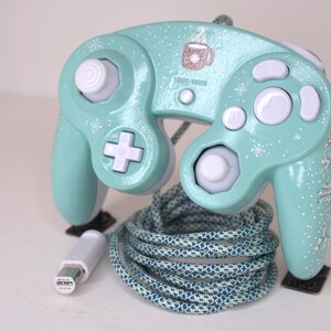 Puede incluir: Un mando de videojuegos con cable en color verde menta, decorado con patrones de copos de nieve blancos y un peque&ntilde;o gr&aacute;fico de una taza de caf&eacute;. Incluye botones blancos, un stick anal&oacute;gico blanco y un cable trenzado con conector blanco.
