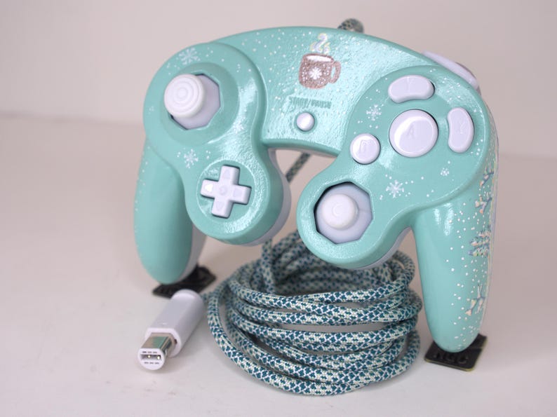 Puede incluir: Un mando de videojuegos con cable en color verde menta, decorado con patrones de copos de nieve blancos y un peque&ntilde;o gr&aacute;fico de una taza de caf&eacute;. Incluye botones blancos, un stick anal&oacute;gico blanco y un cable trenzado con conector blanco.