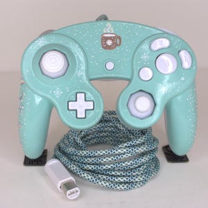 Puede incluir: Un mando de videojuegos color verde menta con un patr&oacute;n de copos de nieve blancos y un gr&aacute;fico de una taza marr&oacute;n con vapor. El mando tiene botones blancos, un joystick anal&oacute;gico blanco y una cruceta blanca. Incluye un cable trenzado azul y blanco.