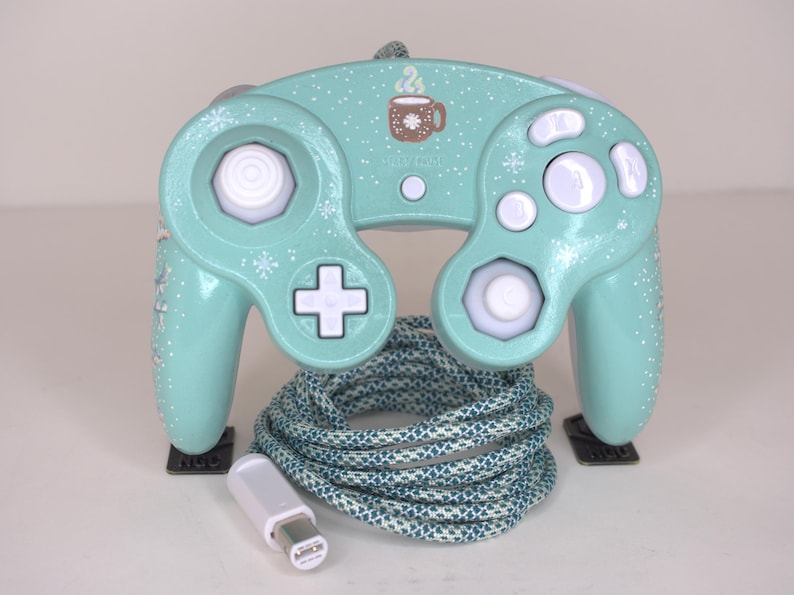 Puede incluir: Un mando de videojuegos color verde menta con un patr&oacute;n de copos de nieve blancos y un gr&aacute;fico de una taza marr&oacute;n con vapor. El mando tiene botones blancos, un joystick anal&oacute;gico blanco y una cruceta blanca. Incluye un cable trenzado azul y blanco.