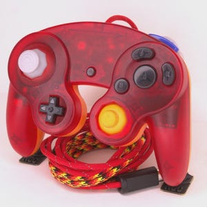 May include: A red translucent video game controller with black buttons, a white joystick, and a yellow and red patterned cord. The controller has a unique design with a distinctive shape and a black connector.