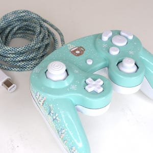 Puede incluir: Un mando de videojuegos de color verde menta con botones, una cruceta y un joystick blancos. El mando est&aacute; decorado con peque&ntilde;os copos de nieve blancos y un gr&aacute;fico de una taza marr&oacute;n. Incluye un cable largo trenzado azul y blanco.