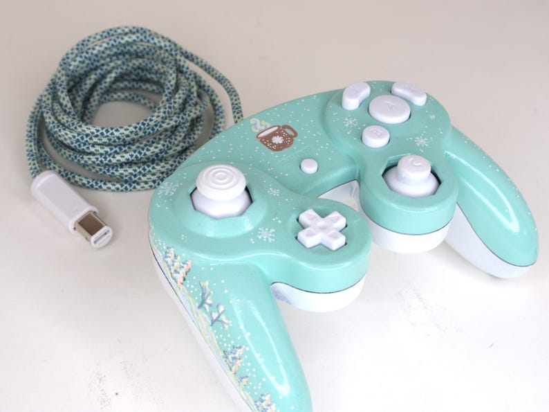 Puede incluir: Un mando de videojuegos de color verde menta con botones, una cruceta y un joystick blancos. El mando est&aacute; decorado con peque&ntilde;os copos de nieve blancos y un gr&aacute;fico de una taza marr&oacute;n. Incluye un cable largo trenzado azul y blanco.