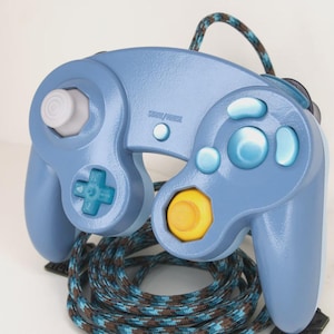 May include: A light blue video game controller with a matching cable. The controller has a gray joystick, a directional pad, and several blue buttons. The central button is yellow. The cable is patterned with blue, brown, and black.